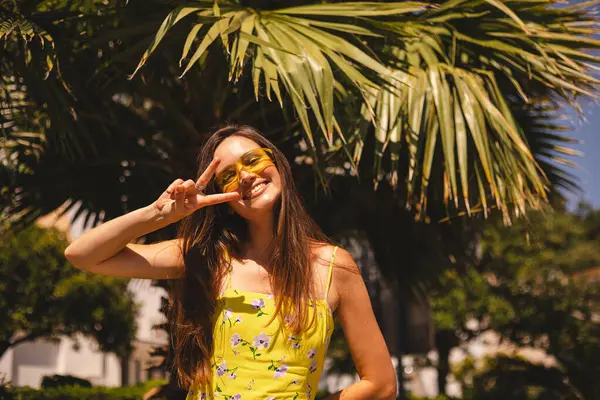 Gülümseyen genç kadın tropik palmiye ağacının önünde barış işareti gösteriyor. Güneşli bir yaz gününde sarı güneş gözlüğü ve çiçekli bir elbise giymek. Neşeli açık hava portresi