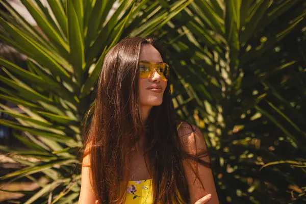 Tropik palmiye ağacının önünde duran gülümseyen genç kadın. Güneşli bir yaz gününde sarı güneş gözlüğü ve çiçekli bir elbise giymek. Neşeli açık hava portresi. Mutlu gülen kız.