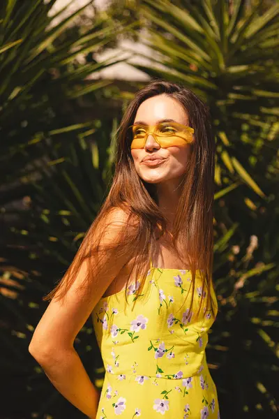 Esmer, uzun saçlı kadın palmiye ağacının yanında duruyor ve sırtına yaslanmış, cilveli bir şekilde gülümsüyor. Sarı elbiseli kadın çiçek desenli ve sarı güneş gözlüklü, yaz havasında..