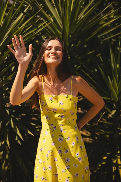 Gülümseyen genç kadın selam veriyor elini tropik palmiye ağacının önünde sallıyor. Güneşli bir yaz gününde sarı çiçekli elbise giymek. Neşeli açık hava portresi.