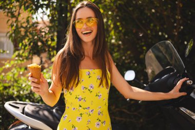 Motorsikletinin yanında duran uzun saçlı mutlu esmer kadın dondurma tutuyor ve gülümsüyor. Kadın parlak sarı elbise ve güneş gözlüğü takıyor, yaz havasında..