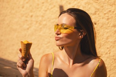Kapalı gözler esmer uzun saçlı kadın sarı güneş gözlüğü takıyor ve elbise giyiyor, havalı görünüyor, gülümsüyor ve eğleniyor, dondurma yiyor. Yaz tatili.