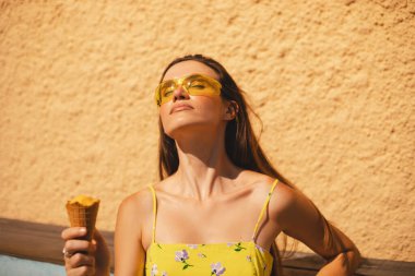 Kapalı gözler esmer uzun saçlı kadın sarı güneş gözlüğü takıyor ve elbise giyiyor, havalı görünüyor, gülümsüyor ve eğleniyor, dondurma yiyor. Yaz tatili.