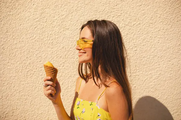 Sıcak ve güneşli bir günde dondurma külahının tadını çıkaran mutlu genç bir kadın. Parlak sarı bir elbise ve güneş gözlüğü takıyor, gülümsüyor ve arka planda bir ışık duvarına yaslanıyor. Yaz yaşam tarzı ve neşesi. Yan görünüm.