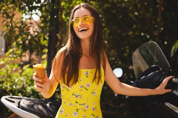 Motorsikletinin yanında duran uzun saçlı mutlu esmer kadın dondurma tutuyor ve gülümsüyor. Kadın parlak sarı elbise ve güneş gözlüğü takıyor, yaz havasında..