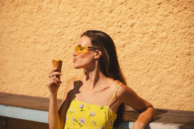 Kapalı gözler esmer uzun saçlı kadın sarı güneş gözlüğü takıyor ve elbise giyiyor, havalı görünüyor, gülümsüyor ve eğleniyor, dondurma yiyor. Yaz tatili.