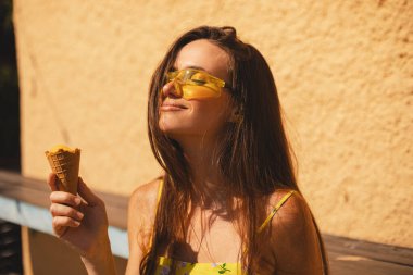 Kapalı gözler esmer uzun saçlı kadın sarı güneş gözlüğü takıyor ve elbise giyiyor, havalı görünüyor, gülümsüyor ve eğleniyor, dondurma yiyor. Yaz tatili.
