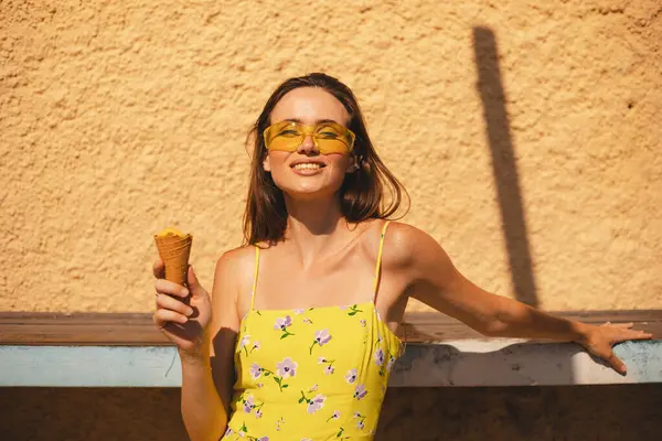 Açık havada waffle külahında lezzetli sarı dondurması olan mutlu genç bayanı kapatın. Sarı güneş gözlüğü takan ve yazın, ilkbaharda ya da güneşli bir günde giyinen bir kız. Esmer kadın gülüyor..