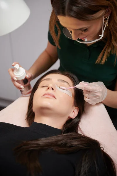 Close up of beauty specialist in sterile gloves applying cleansing mousse on woman eyelashes with cosmetic lash brush. Eyelash stylist preparing woman lashes for applying extensions.