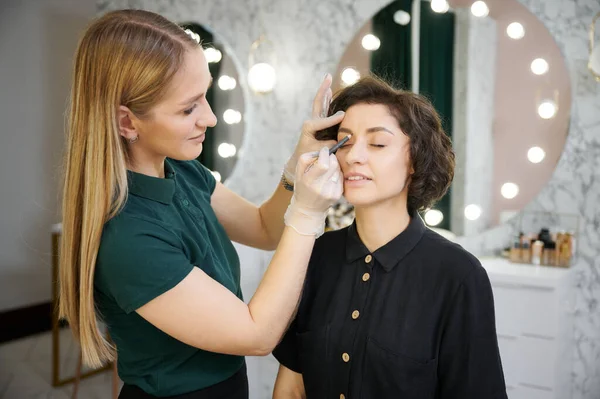 Young woman with closed eyes having eyebrow correction procedure in beauty salon. Female makeup artist using eyebrow pencil while correcting client brows in visage studio.
