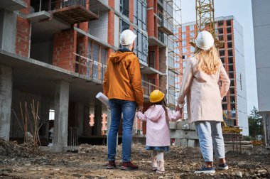 Güvenlik kasklı bir kadın ve erkeğin, kızlarının elini tutarken ve inşaat alanındaki vinç kulesine bakarken. Dışarıda inşaat halinde olan çocuklu bir aile..