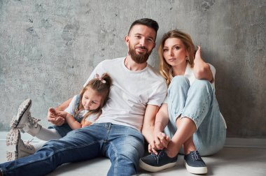 Sevgi dolu ebeveynler ve yeni dairede duvarın kenarında oturan küçük bir çocuk. Kadın ve erkek el ele tutuşup kameraya bakarken yeni evinde kızına sarılıyorlar..