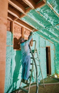 Male builder insulating wooden frame house. Man worker spraying polyurethane foam inside of future cottage, standing on ladder, using plural component gun. Construction and insulation concept.