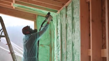 Male builder insulating wooden frame house. Man worker spraying polyurethane foam inside of future cottage, using plural component gun. Construction and insulation concept.