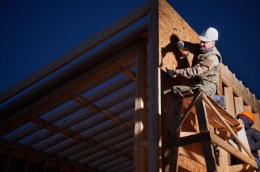Carpenters hammering nail into OSB panel on the wall of future cottage in the evening. Men workers building wooden frame house. Carpentry and construction concept.
