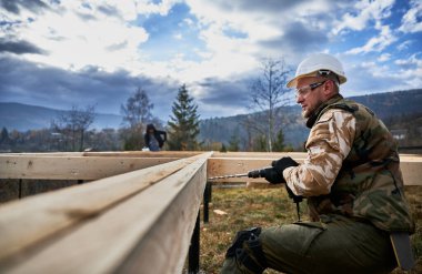 Yığınla temele ev inşa eden bir adam. Erkek işçi, gelecekteki evin ahşap çerçevesinde elektrikli matkapla delik açıyor. Beyaz kask takan ve koruyucu gözlük takan bir inşaatçı. Marangozluk kavramı.