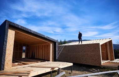 Inspector building wooden frame house on pile foundation in Scandinavian style barnhouse. Bald man standing on construction site, inspecting quality of work on sunny day with blue sky on background.