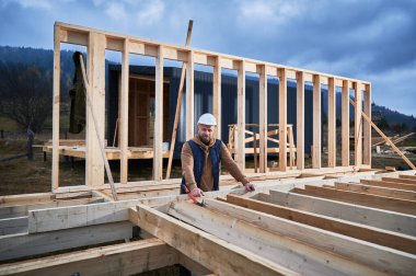 Erkek mimar ahşap çerçeve inşa ediyor. İnşaat alanında güvenlik kaskıyla duran adam, iş kalitesini denetliyor..
