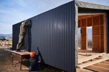 Male builders installing black corrugated iron sheet used as facade of future cottage. Men workers building wooden frame house in the Scandinavian style barnhouse. Carpentry and construction concept.