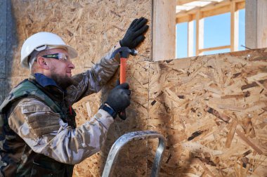 Marangoz, OSB panelinin duvarına çivi çakıyor. Ahşap çerçeve inşa eden bir işçi. Marangozluk ve inşaat konsepti.