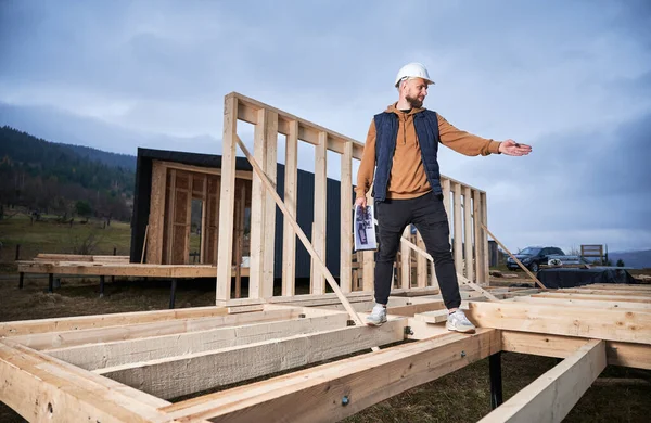 stock image Male engineer building wooden frame house. Man builder standing on construction site in safety helmet, with construction documentation inspecting quality of work.