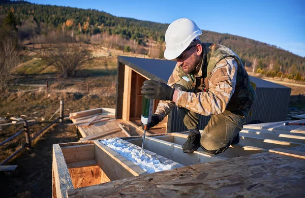 Male builder doing thermal insulation on roof of wooden frame house. Man worker spraying polyurethane foam on rooftop of future cottage. Construction and insulation concept.
