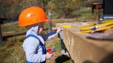 Boy toddler playing as builder on construction site. Child carpenter in orange helmet and blue overalls learning to build wooden frame house outdoor on sunny day. Carpentry and workshop concept.
