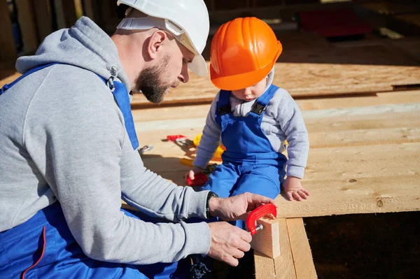 Father with toddler son building wooden frame house. Male builder learing kid to use clamp on construction site, wearing helmet and blue overalls on sunny day. Carpentry and family concept.