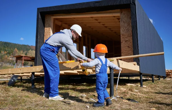 Father with toddler son building wooden frame house. Male builder and kid playing with tape measure on construction site, wearing helmet and blue overalls on sunny day. Carpentry and family concept.