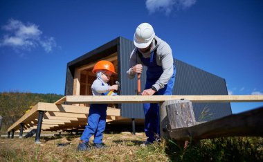 Father with toddler son building wooden frame house. Male builders hammering nail into plank on construction site, wearing helmet and blue overalls on sunny day. Carpentry and family concept.