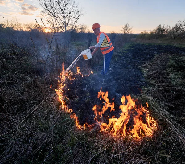 stock image Fireman ecologist fighting wildfire in field with evening sky on background. Male firefighter pouring water on burning dry grass near yellow triangle with skull and crossbones warning sign.