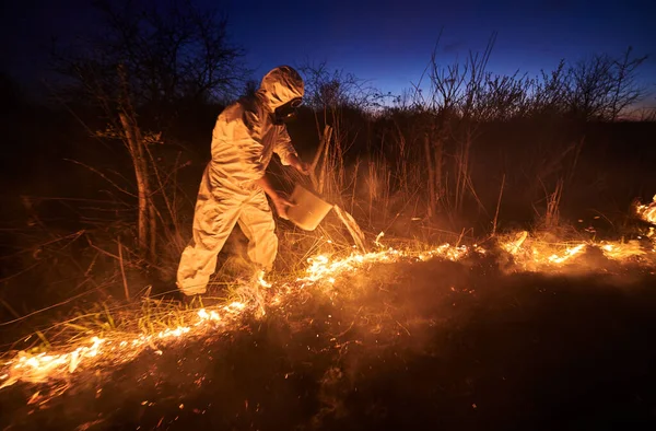 Firefighter extinguishing fire in field with blue night sky on background. Man in protective radiation suit and gas mask holding bucket and pouring water on burning grass. Natural disaster concept.