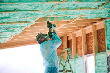 Male builder insulating wooden frame house. Man worker spraying polyurethane foam inside of future cottage, using plural component gun. Construction and insulation concept.