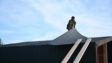 Male builder installing black corrugated iron sheet used as facade of future cottage. Man worker building wooden frame house. Carpentry and construction concept.