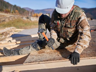 Marangoz, gelecekteki kulübenin çatısında OSB paneline çivi çakıyor. Ahşap çerçeve inşa eden bir işçi. Marangozluk ve inşaat konsepti.