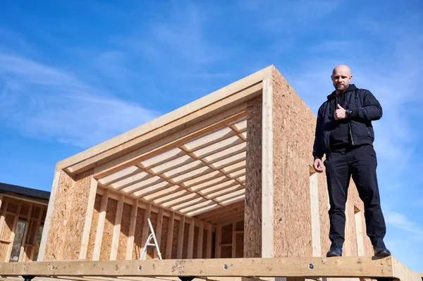 Developer building wooden frame house in Scandinavian style barnhouse. Bald man standing on construction site, inspecting quality of work, showing thumbs up on sunny day with blue sky on background.