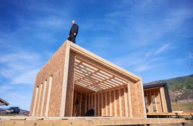Male builder building wooden frame house. Bald man standing on construction site, inspecting quality of work on sunny day with blue sky on background.
