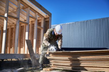 Carpenter using circular saw for cutting wooden OSB board. Man worker building wooden frame house on pile foundation. Carpentry concept.