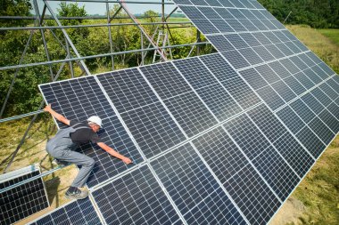 Güneşli bir günde güneş panelleri kuran bir işçi. Kask ve üniforma giyen Avrupalı bir adam. Modüllerin PV panel olarak foto-voltaik koleksiyonu. Çevre güvenliği fikri. Yeşil enerji.