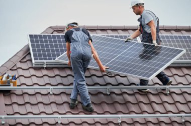 Erkek teknisyenler evin çatısında fotovoltaik güneş modülü taşıyorlar. Kasklı işçiler dışarıda güneş paneli sistemi kuruyor. Alternatif ve yenilenebilir enerji kavramı.
