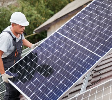 Evin çatısında fotovoltaik güneş modülü taşıyan teknisyen. Kasklı elektrikçi dışarıda güneş paneli kuruyor. Alternatif ve yenilenebilir enerji kavramı.