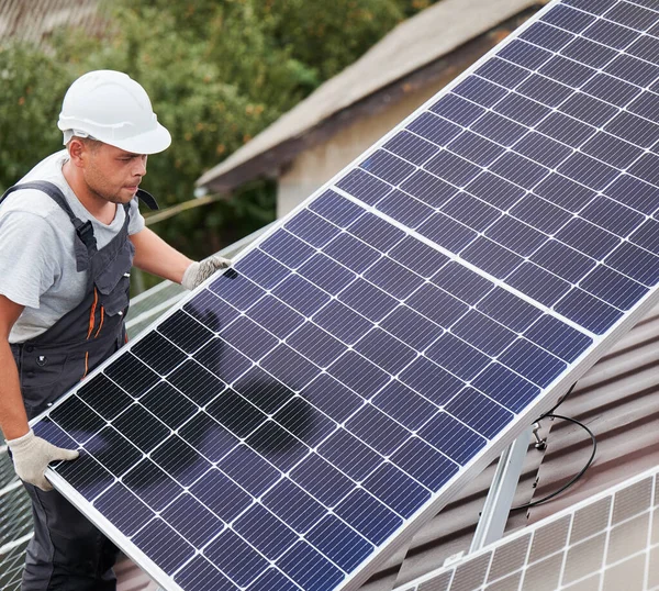 屋根の上に太陽光発電モジュールを運ぶ男技術者 太陽電池パネルシステムを屋外に設置ヘルメットの電気技師 再生可能エネルギーの概念 — ストック写真