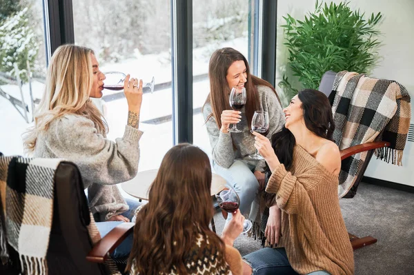 Young women enjoying winter weekends inside contemporary barn house. Four girls having fun and drinking red wine.