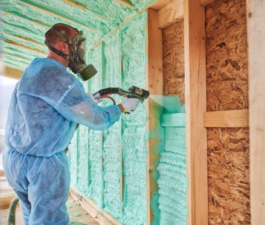 Male builder insulating wooden frame house. Man worker spraying polyurethane foam inside of future cottage, using plural component gun. Construction and insulation concept.