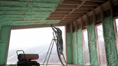 Male builder insulating wooden frame house. Man worker spraying polyurethane foam inside of future cottage, using plural component gun. Construction and insulation concept.