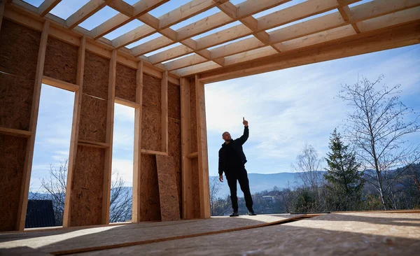 Male developer building wooden frame house in the Scandinavian style barnhouse. Bald man standing on construction site, inspecting quality of work on sunny day with blue sky on background.