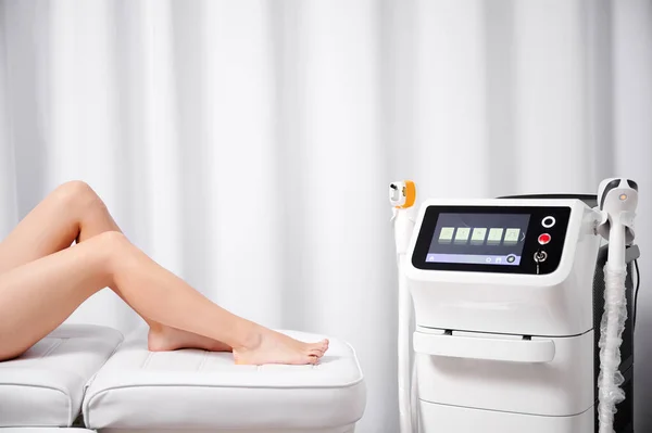 Close up of young woman sitting on daybed near diode laser hair removal machine in cosmetology clinic. Beautiful female legs and diode laser epilation machine in beauty salon.