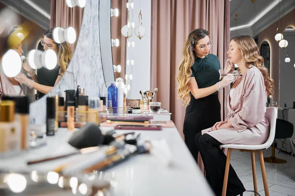 Female beauty specialist applying foundation with cosmetic brush. Young woman sitting at dressing table while stylist in sterile gloves doing professional makeup in visage studio.