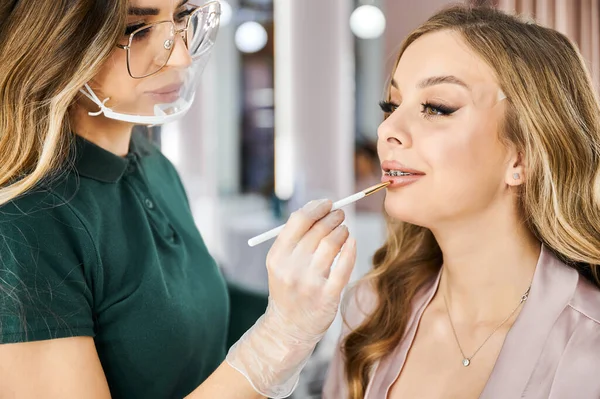Close up of female beauty specialist in protective mask applying lipstick on woman lips with cosmetic lip brush. Makeup artist in face mask doing professional makeup for client with braces on teeth.