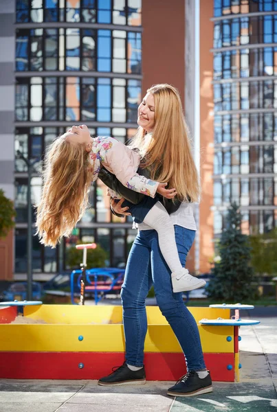 Female holding adorable daughter upside down at playground. Girl having fun with attractive blond mother at modern courtyard of city residential high-rise buildings. Concept of family and childhood.